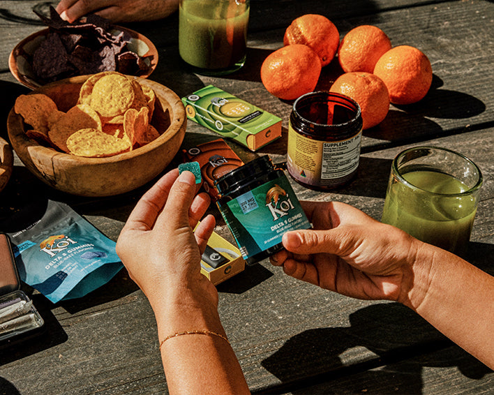Two hands holding Koi gummies on a table with snacks and drinks.