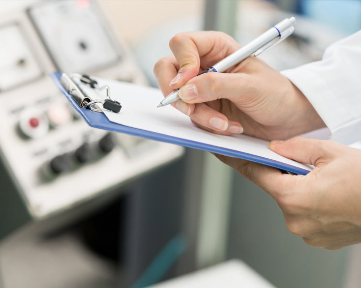 Person holding a pen and writing on a clipboard with medical equipment in the background